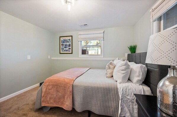 A bedroom with a bed, a window with blinds, a wall-mounted shelf with a plant, and a decorative lamp.