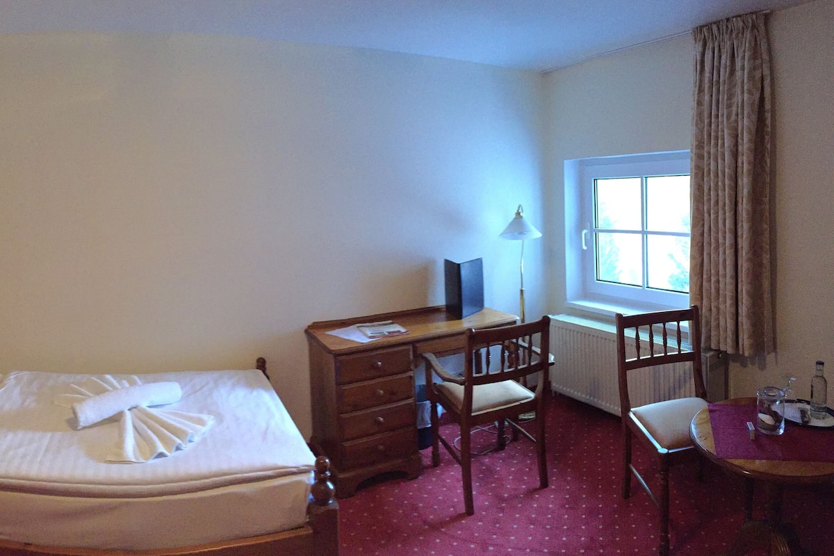 Quarto de hotel com cama bem arrumada, penteadeira de madeira, escrivaninha com cadeira e janela com cortinas.