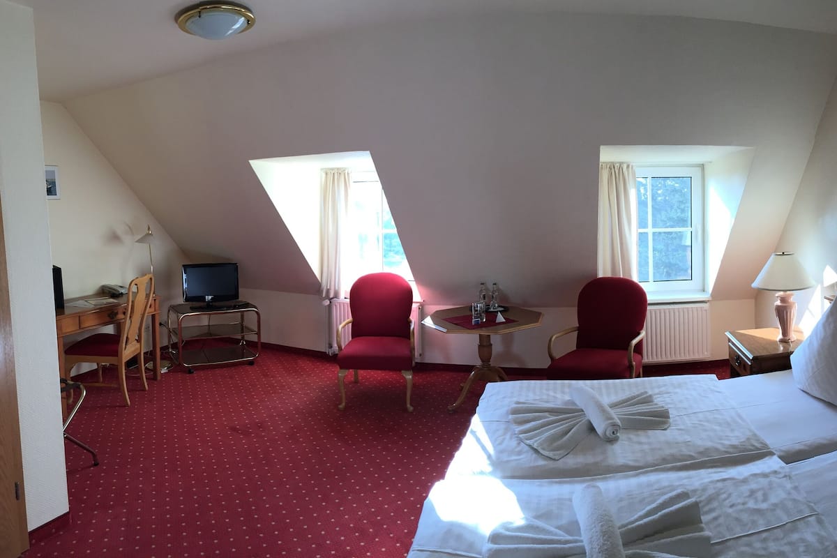 Quarto de hotel com uma cama, duas cadeiras vermelhas, uma mesinha com abajur, uma escrivaninha com computador e um quadro na parede.