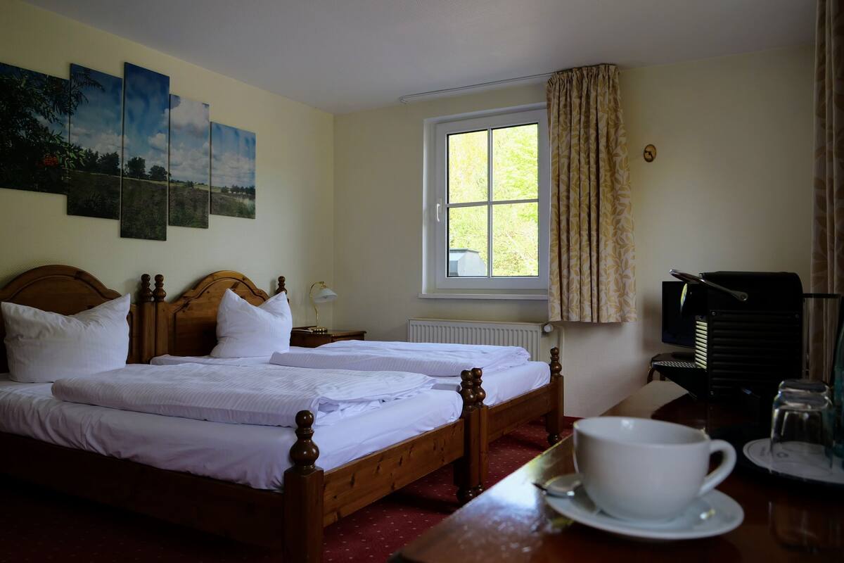 Quarto de hotel com duas camas, uma janela com cortinas, uma mesa com xícara e pires, e uma televisão.