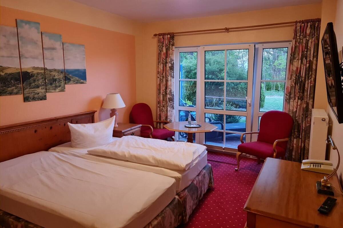 Quarto de hotel com uma cama grande, duas cadeiras, uma mesinha e uma janela com cortinas.