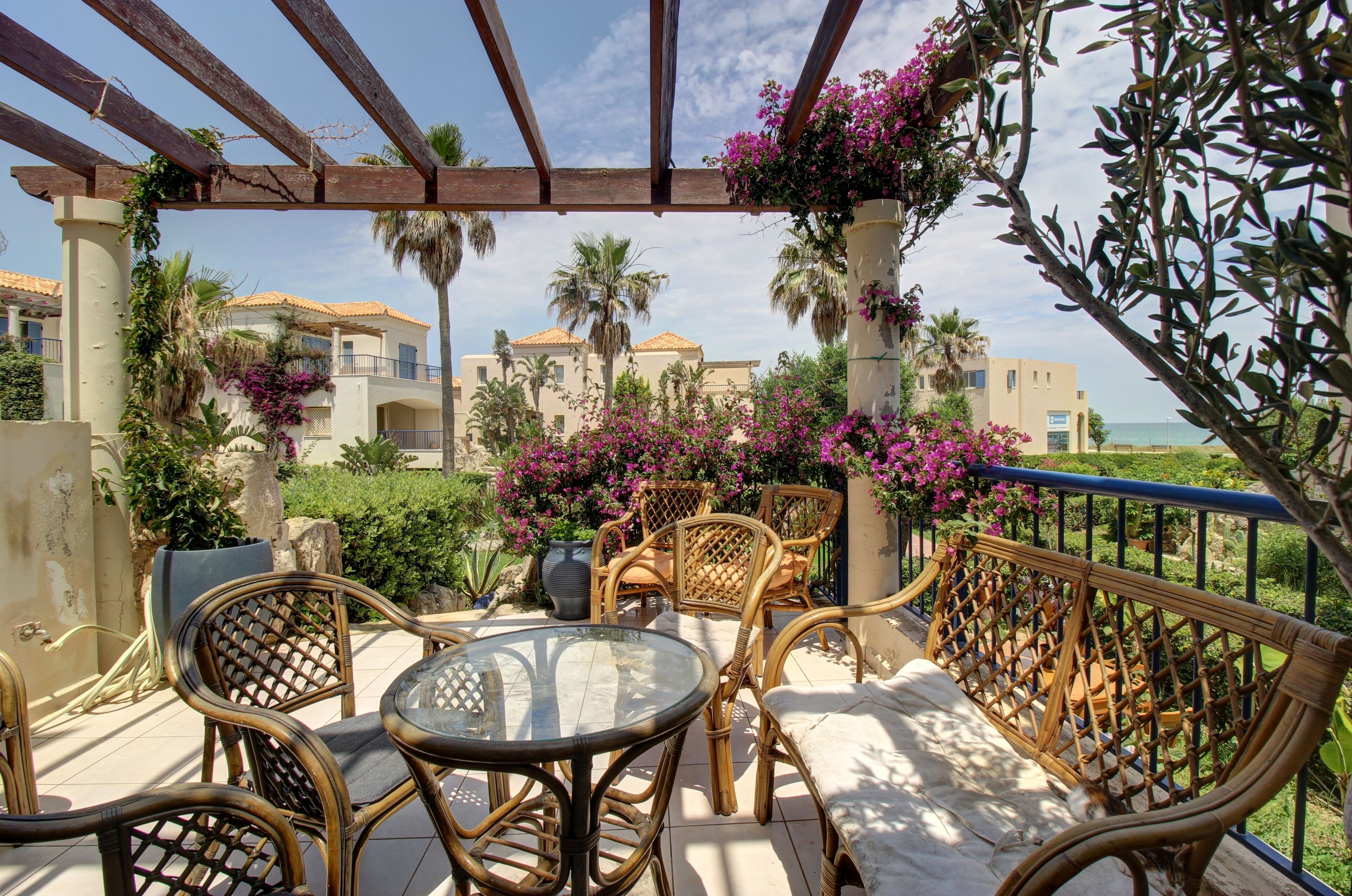 Un espacio de estar al aire libre techado con mobiliario de mimbre y una mesa de vidrio, con vistas a un jardín ajardinado con flores púrpuras y palmeras.