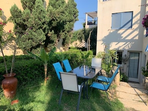 An outdoor patio with a table and chairs, surrounded by greenery and a building with a window. - 3 Bedroom Apartment Elias Total (Platanias)