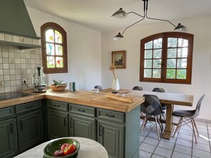 Une cuisine avec des meubles de rangement verts, un plan de travail en bois, une table à manger avec des chaises et deux fenêtres ornées de vitraux.