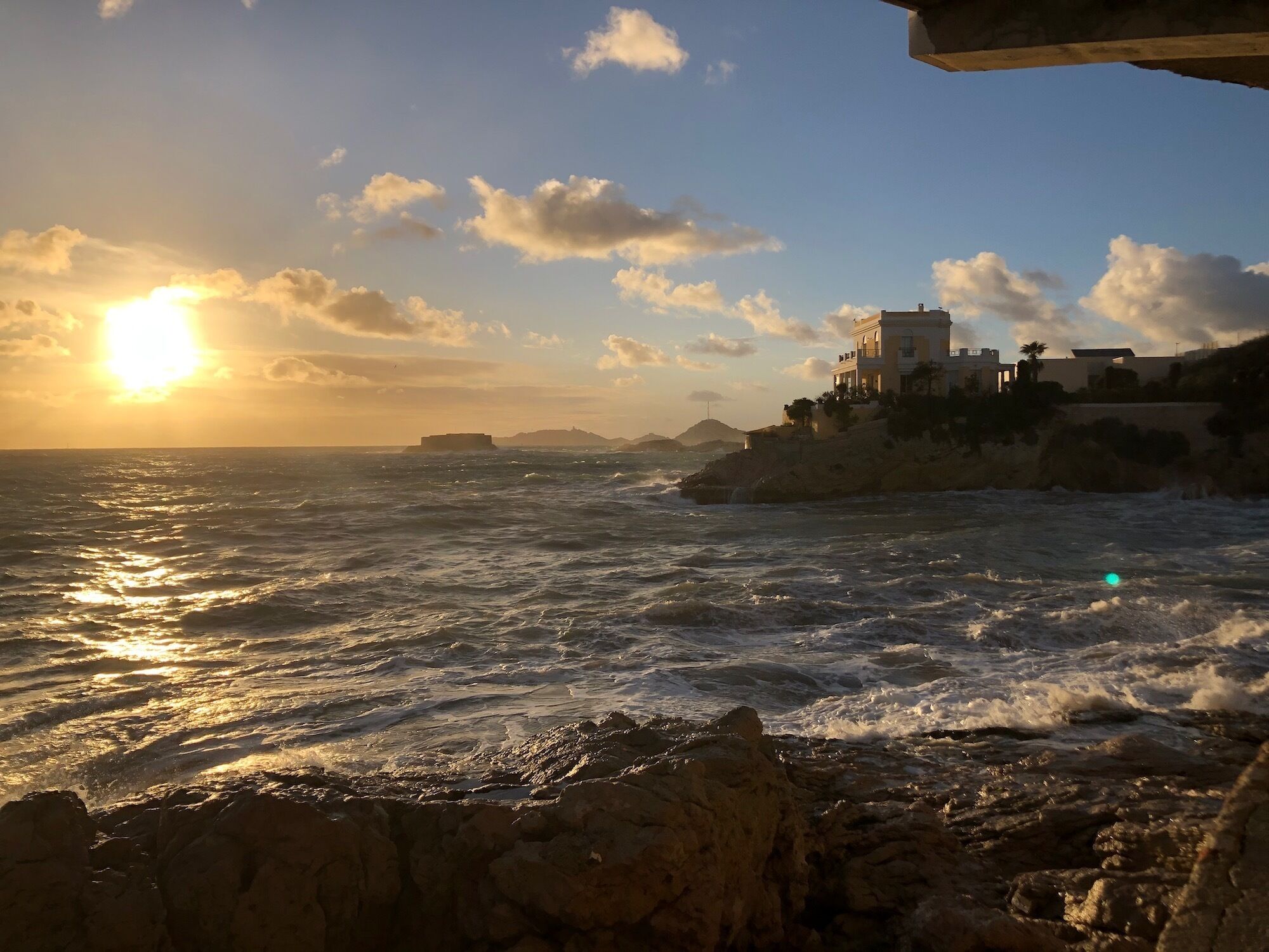 A coastal scene with a building on a rocky outcrop, waves crashing against the shore, and the sun setting in the background.