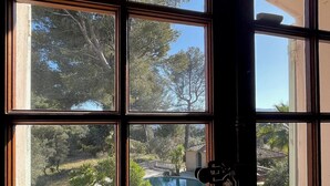 A view through a window with a pool, trees, and a building in the background. - Villa Grand Cédre - Family house with infinity pool, garden 4000m2. (Marseille)