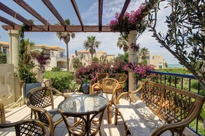 A covered outdoor seating area with wicker furniture and a glass table, overlooking a landscaped garden with purple flowers and palm trees. - 1 Bedroom Apartment on Beach Elias Upper (Platanias)