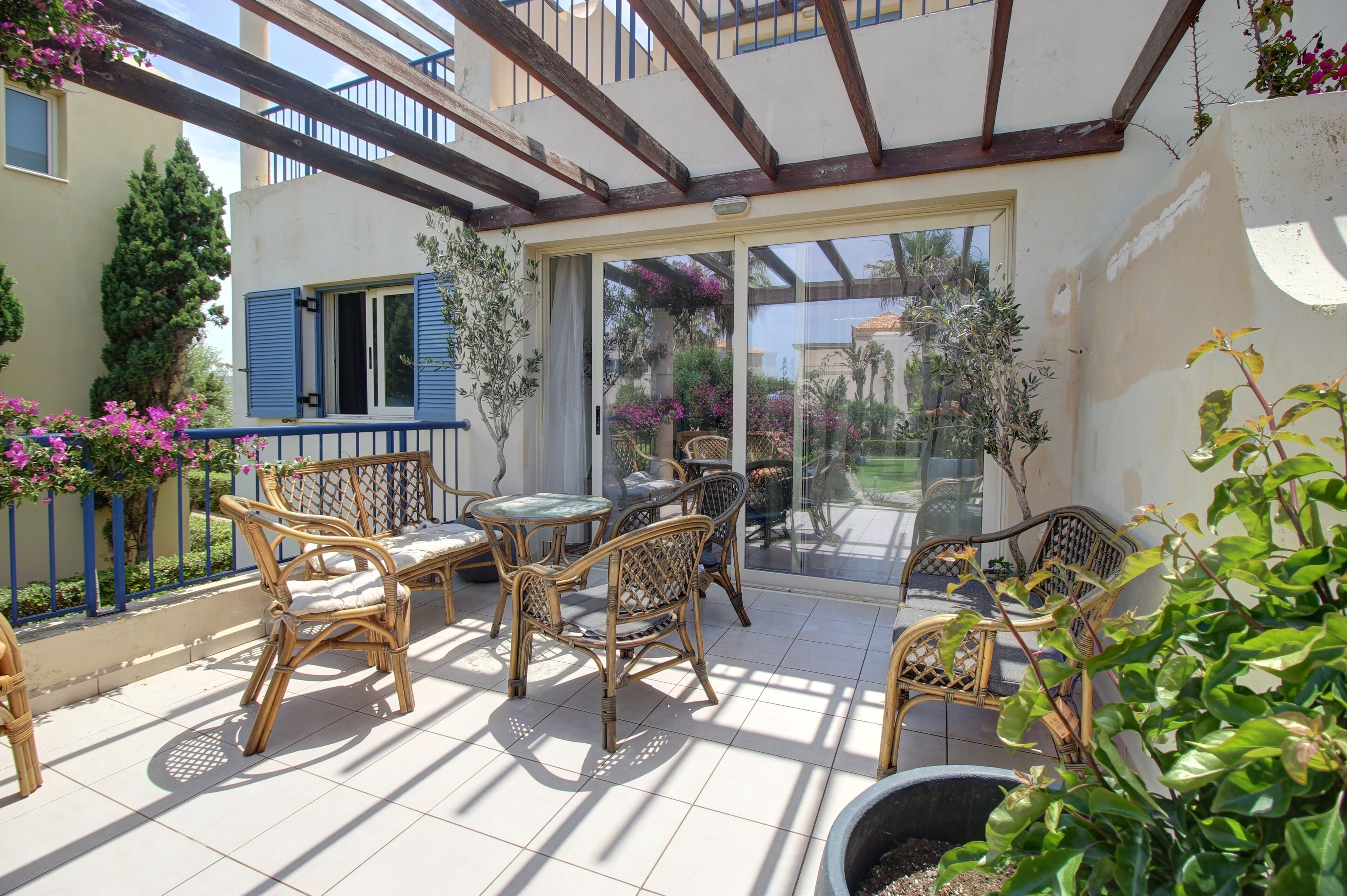 Een overdekt terras met rieten meubilair, een glazen deur naar een tuin en een blauwe balustrade.