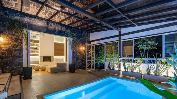 Un área de piscina moderna al aire libre con un muro de piedra, una zona de estar y un ventanal que da a una habitación con chimenea.