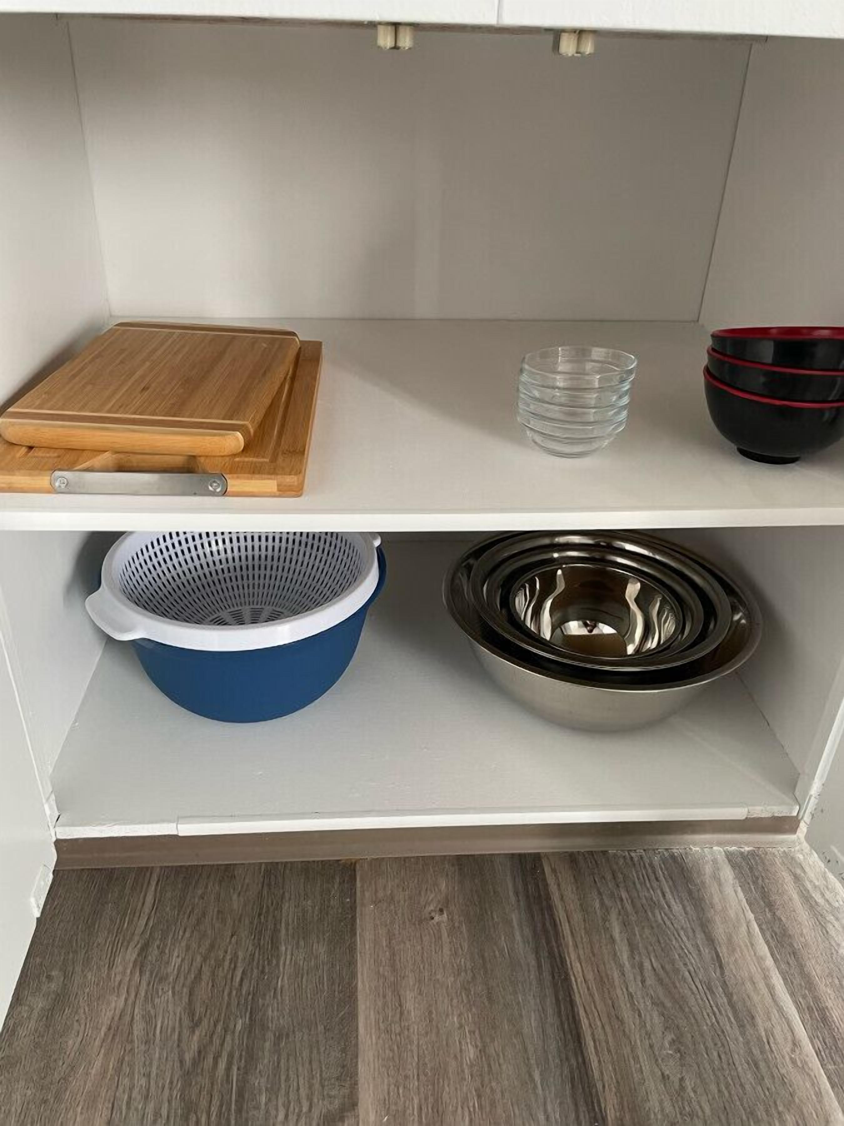 A kitchen with a wooden cutting board, a colander, and a metal bowl on the upper shelf, and a blue colander and a metal bowl on the lower shelf.
