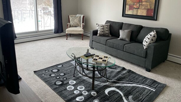 Una sala de estar con un sofá gris, una mesa de centro de cristal, un tapiz estampado, una silla y un cuadro en la pared.
