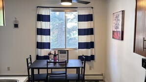 Une cuisine avec une table à manger, des chaises, un ventilateur de plafond, une fenêtre avec des rideaux et un tableau au mur.