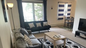 Un salon avec un canapé, une table basse et un téléviseur. Il y a une fenêtre avec des rideaux et une vue sur l’extérieur.