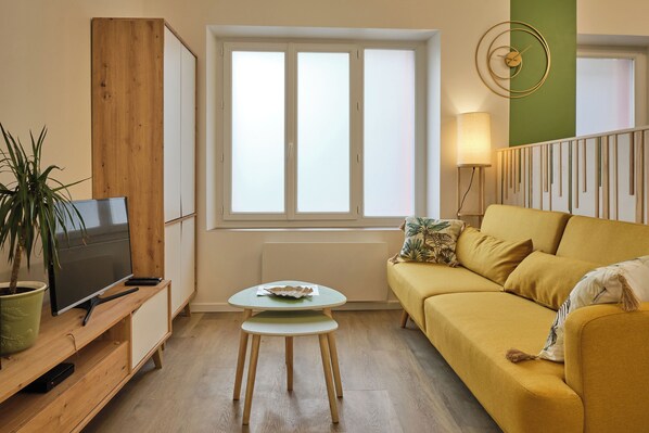 Una sala de estar con un sofá, una mesita, un televisor sobre un mueble de madera, una ventana con persianas y una lámpara.
