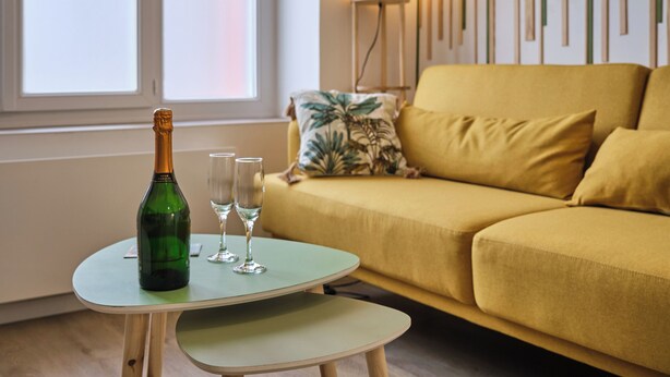 A green bottle of champagne, two glasses, and a small table in a room with a yellow sofa and a window.