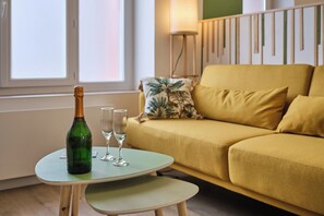 Una botella de champán verde, dos copas y una mesita en una habitación con un sofá amarillo y una ventana.
