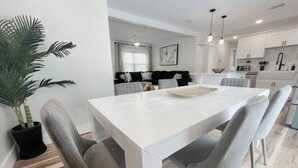 A modern dining area with a white table and grey chairs, a potted plant, and a kitchen in the background.