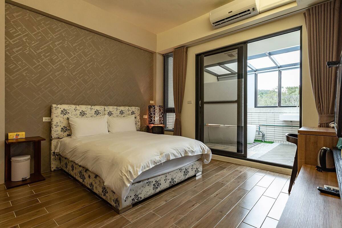 Habitación de hotel con cama, mesita de noche, un cubo de basura, escritorio con silla y ventana con cortinas.