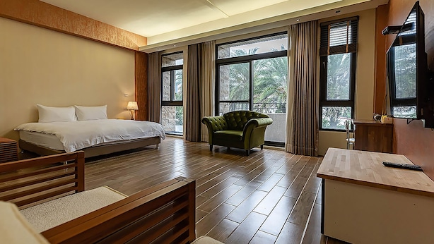 Una habitación de hotel con una cama, un sillón verde, un escritorio y ventanas amplias.