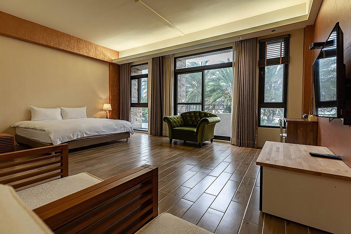 Una habitación de hotel con una cama, un sillón verde, un escritorio y ventanas amplias.