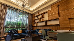Una sala de estar moderna con un sofá modular, una mesa de centro y un escritorio con una silla. Hay un ventanal con cortinas, una araña y estanterías empotradas de madera.
