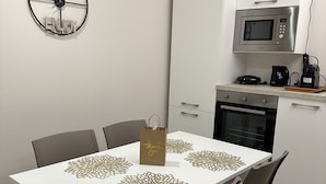Un comedor con una mesa blanca, cuatro sillas y posavasos decorativos. Se ve un reloj y un microondas al fondo.