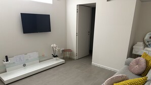 Una sala moderna con una televisión de pantalla plana, una mesa de centro de vidrio, un sofá con cojines y una puerta que da a otra habitación.