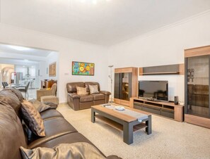 A modern living room with a leather sofa, a wooden coffee table, a TV, and a dining area in the background.