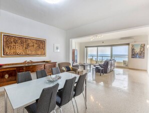 A spacious living area with a dining table, chairs, and a large window offering a view of the sea.