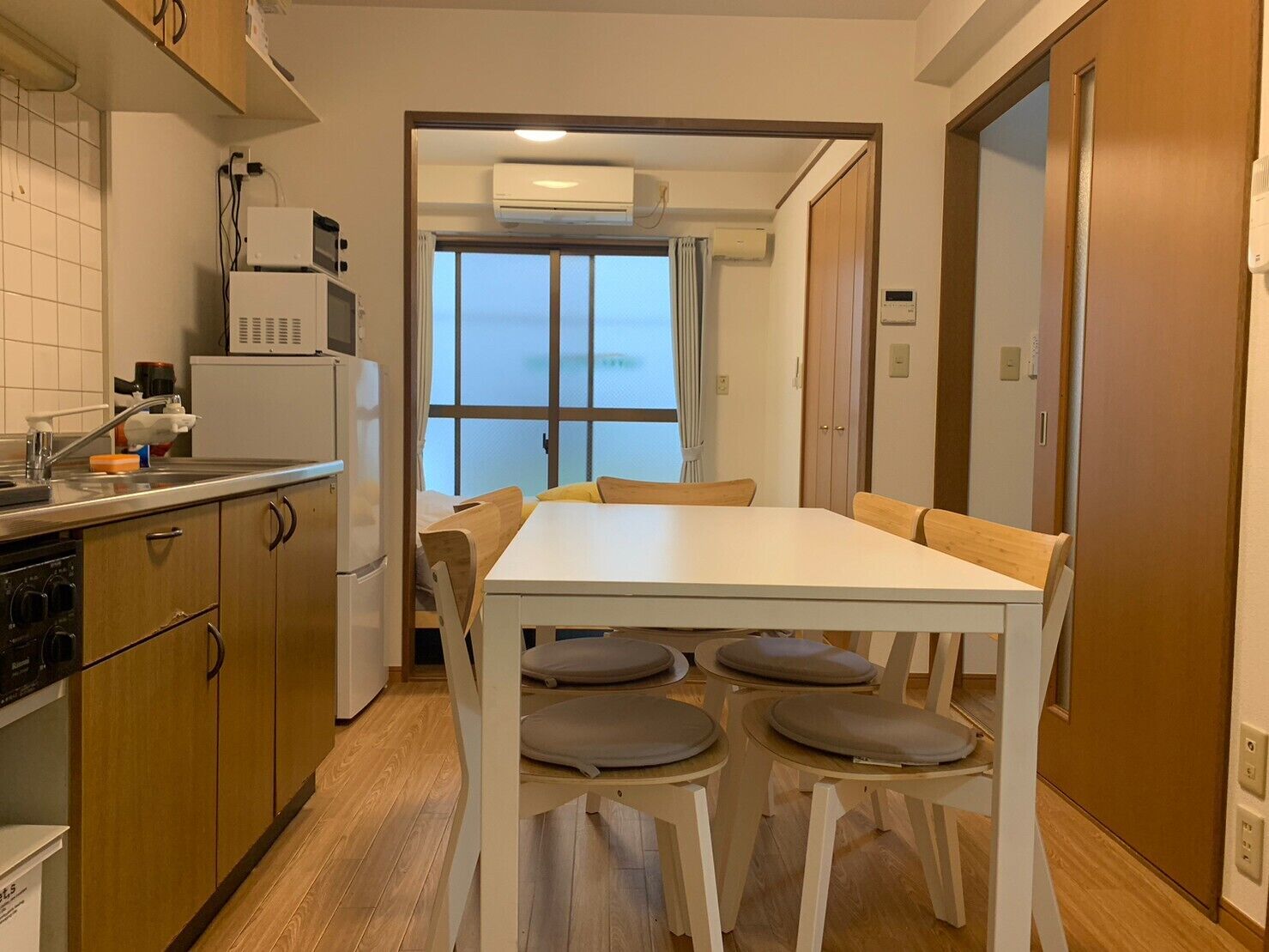 Une cuisine compacte avec un coin repas, comprenant une table et des chaises blanches, des meubles de rangement en bois et une fenêtre avec des rideaux.
