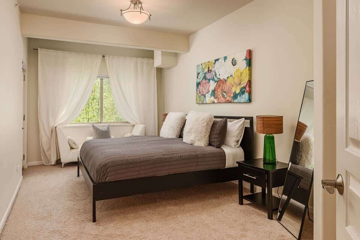 A bedroom with a bed, a nightstand, a lamp, a painting, and a window with curtains.
