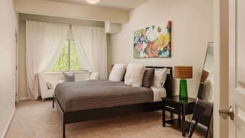 A bedroom with a bed, a nightstand, a lamp, a painting, and a window with curtains.