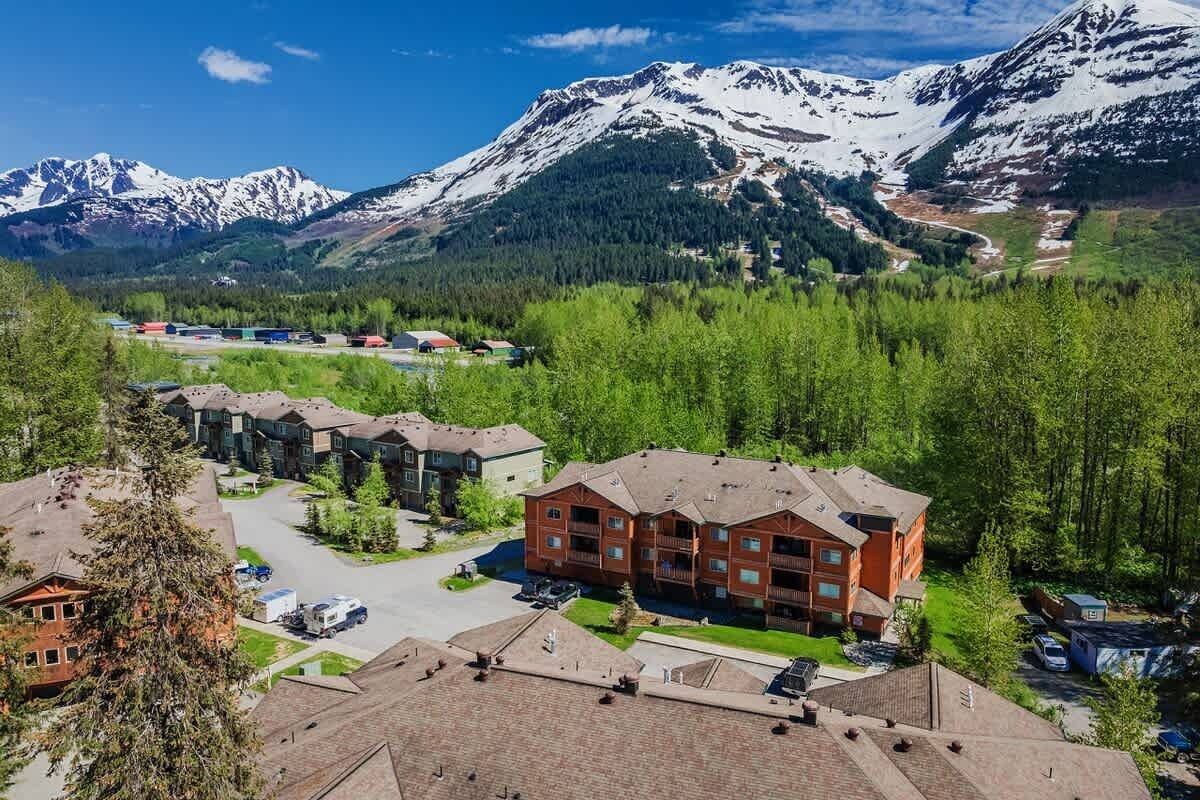 A multi-story building with a parking lot in front, surrounded by trees and mountains in the background.