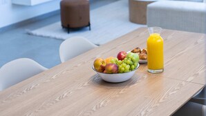 A modern living room with a wooden dining table, a bowl of fruit, and a glass of orange juice. A flat-screen TV is mounted on the wall, and there is a sofa and a coffee table in the background. - SeaFront Luxurious  3BR Apartment | Mamad (Rishon LeTsiyon)
