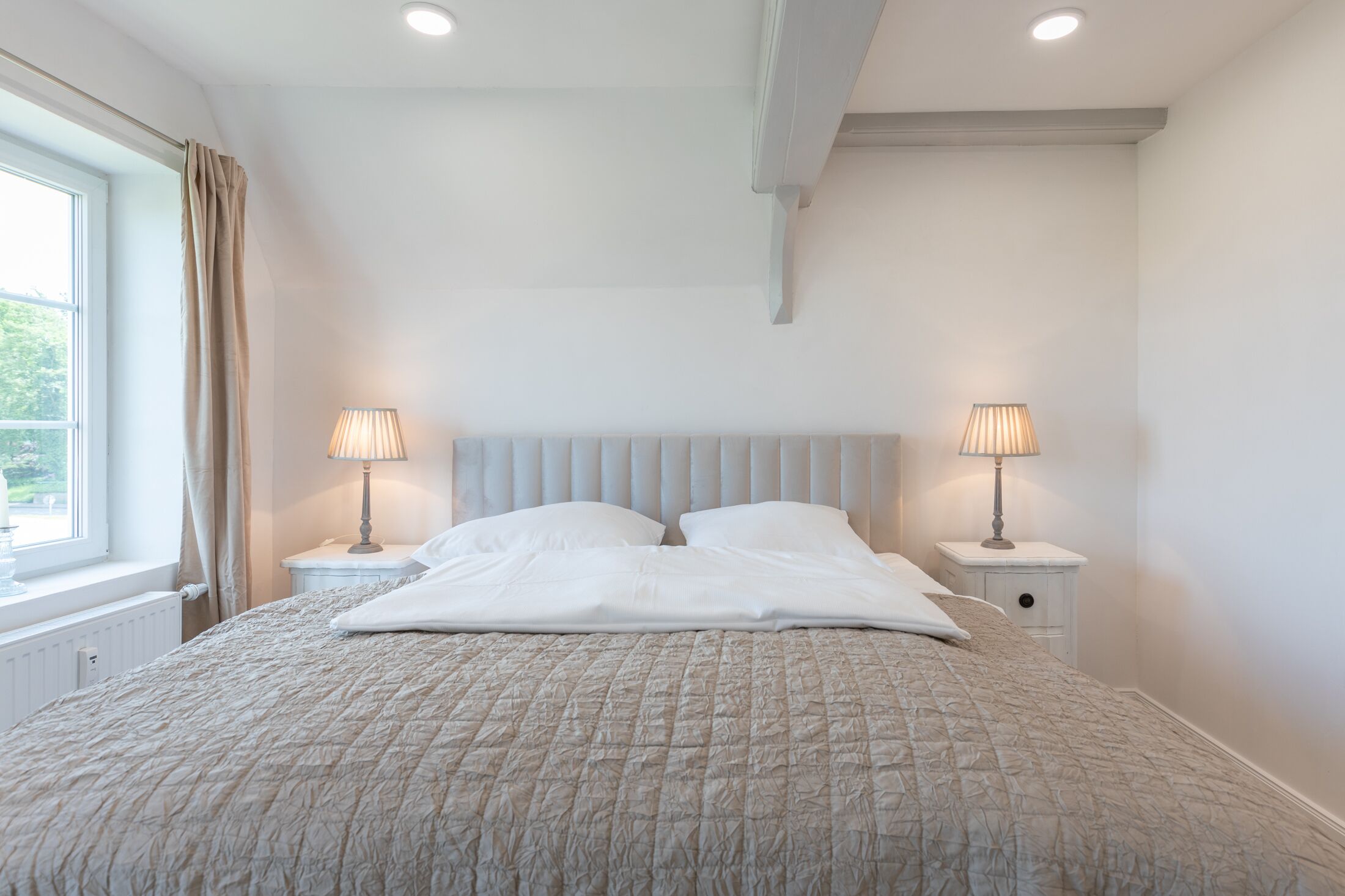 A bedroom with a large bed, two bedside tables with lamps, a window with curtains, and a radiator.