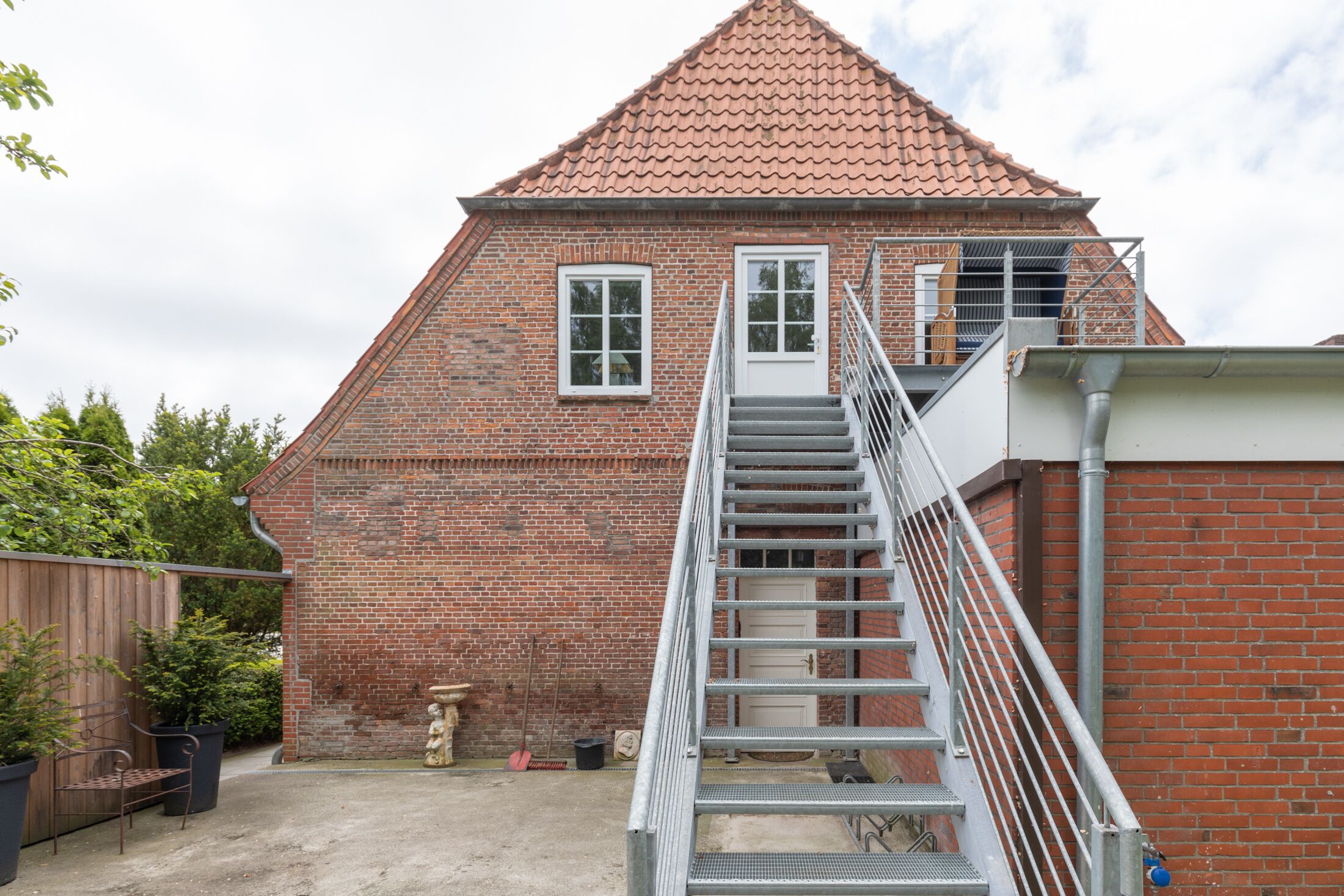 A brick building with a red-tiled roof, a staircase leading to a door, and a fenced garden area.