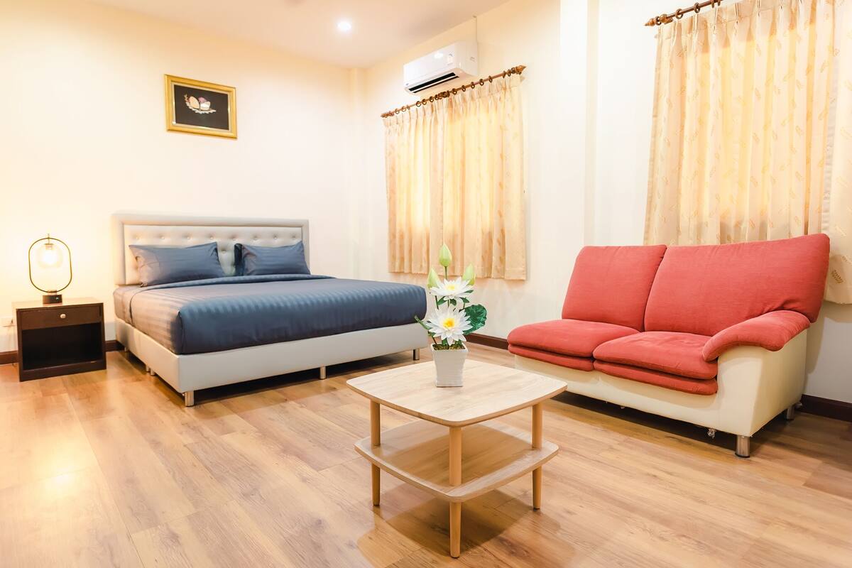 A bedroom with a bed, a red sofa, a small table with a flower vase, and a nightstand with a lamp.