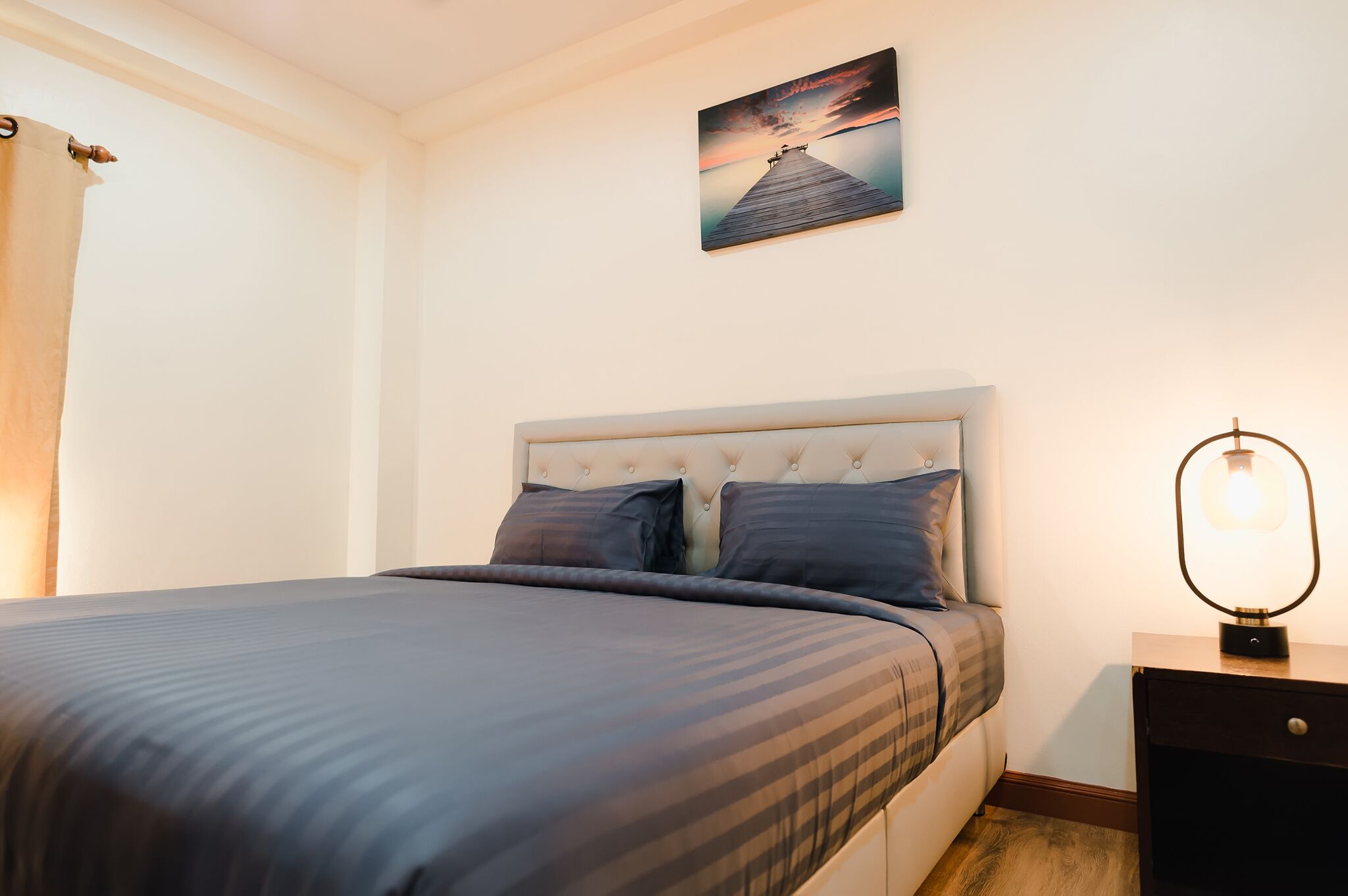 A neatly made bed with a headboard, a bedside table with a lamp, and a painting on the wall.