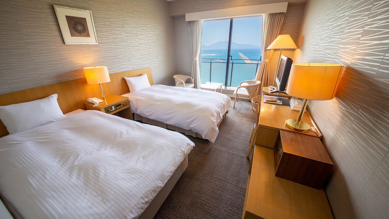 Une chambre d’hôtel avec deux lits, un bureau avec une lampe, une chaise, une petite table et une vue sur la mer.