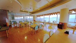A spacious dining area with wooden floors and tables, large windows with a view of the sea, and a high ceiling with lighting fixtures. - Kokumin Shukusya Marin Terrace Ashiya (Ashiya)