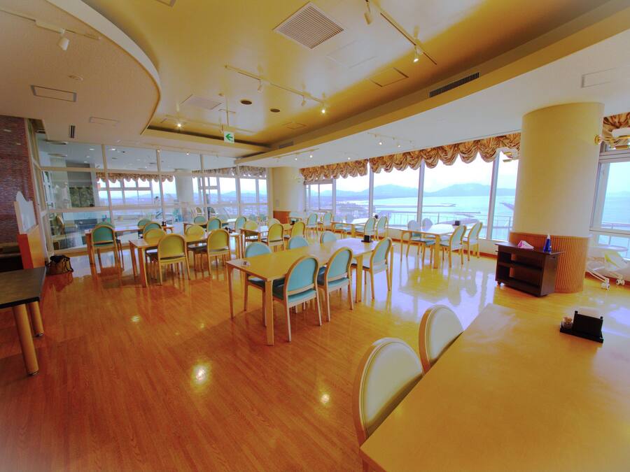 A spacious dining area with wooden floors and tables, large windows with a view of the sea, and a high ceiling with lighting fixtures.