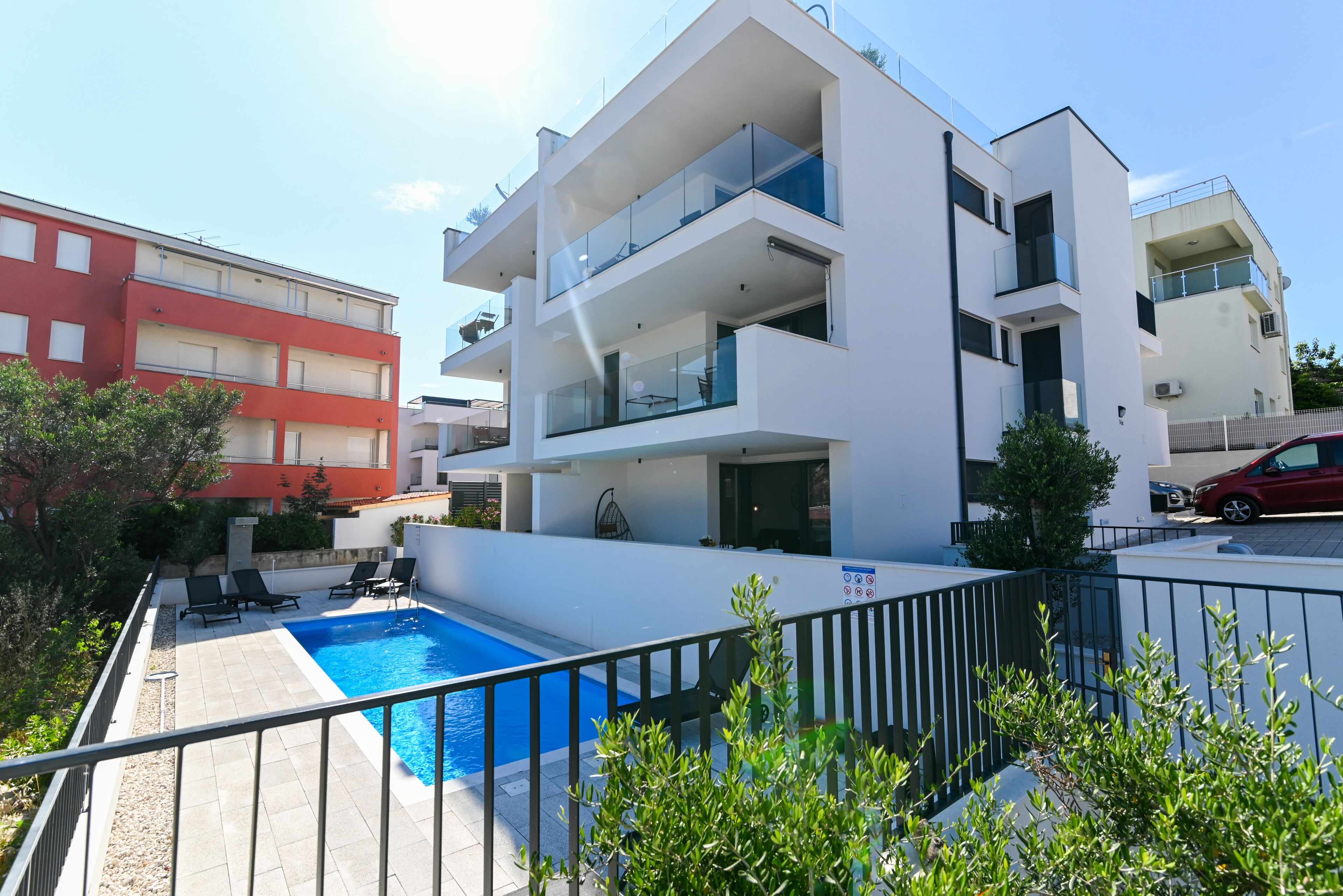 Un moderno edificio de apartamentos con piscina, balcones y un edificio rojo al fondo.