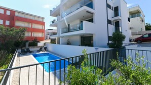 Un moderno edificio de apartamentos con piscina, balcones y un edificio rojo al fondo.