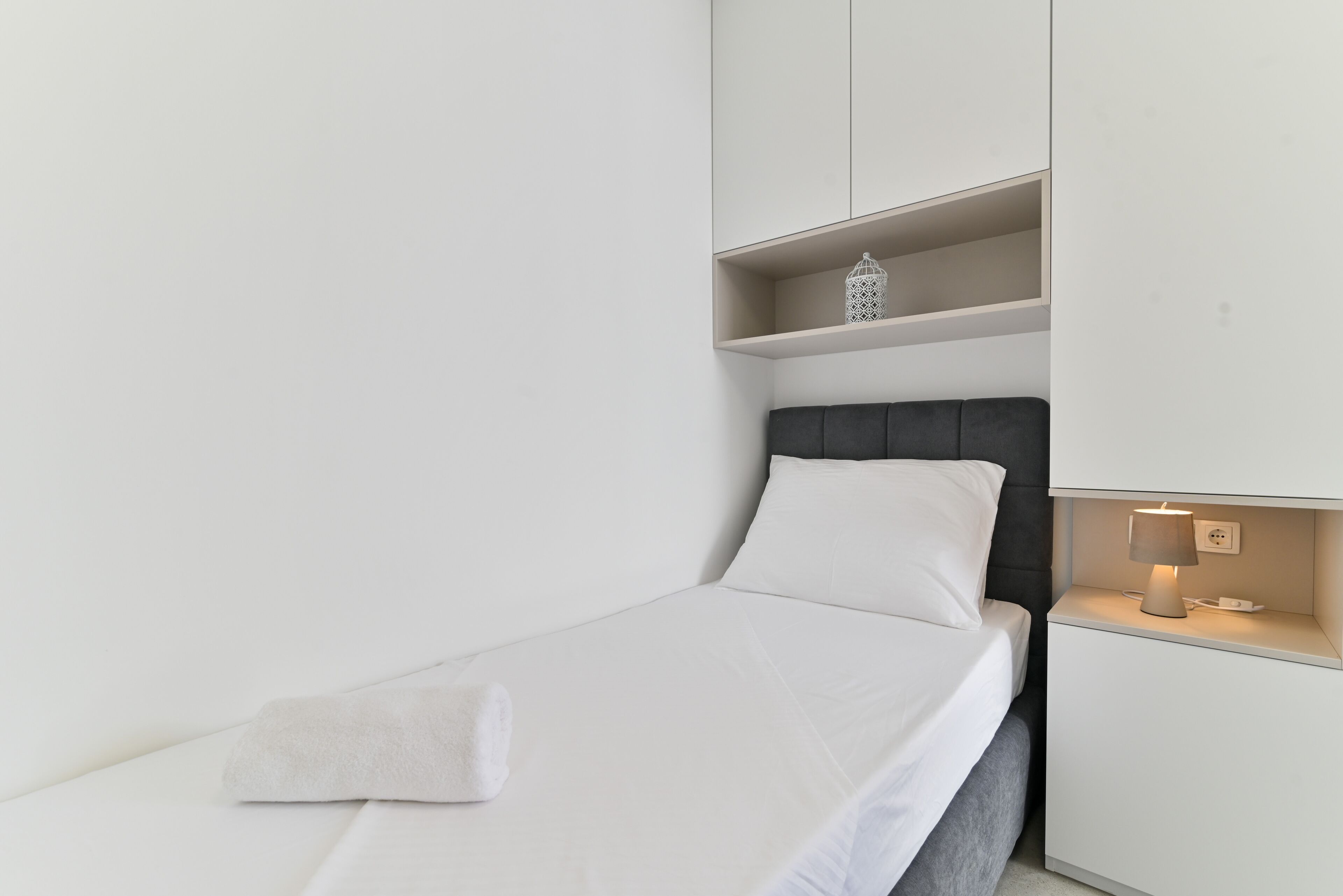 Un lit bien fait, avec un drap blanc et une serviette pliée, une lampe de chevet et un petit objet décoratif posé sur une étagère au-dessus du lit.