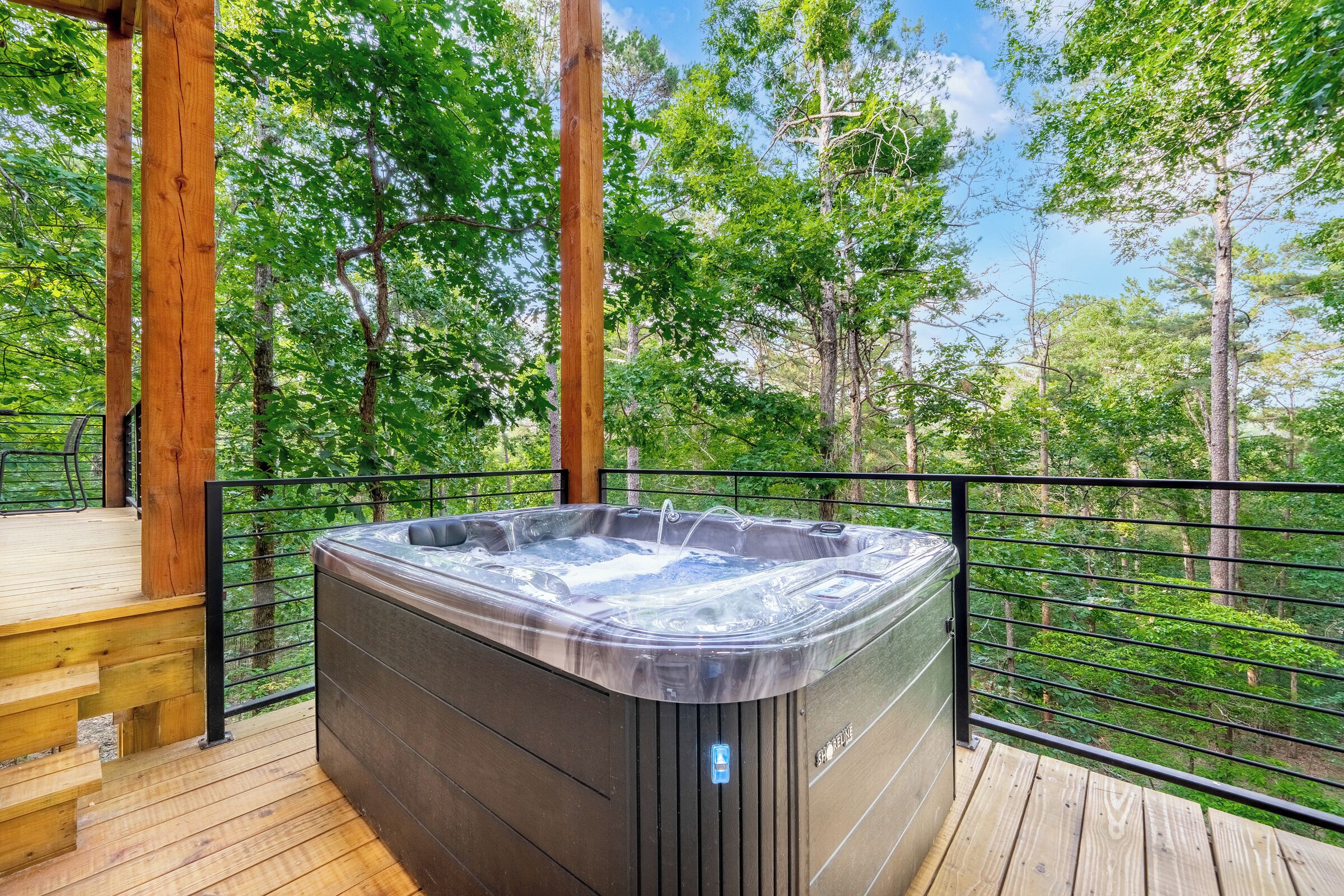 A hot tub on a wooden deck with a metal railing, surrounded by trees.