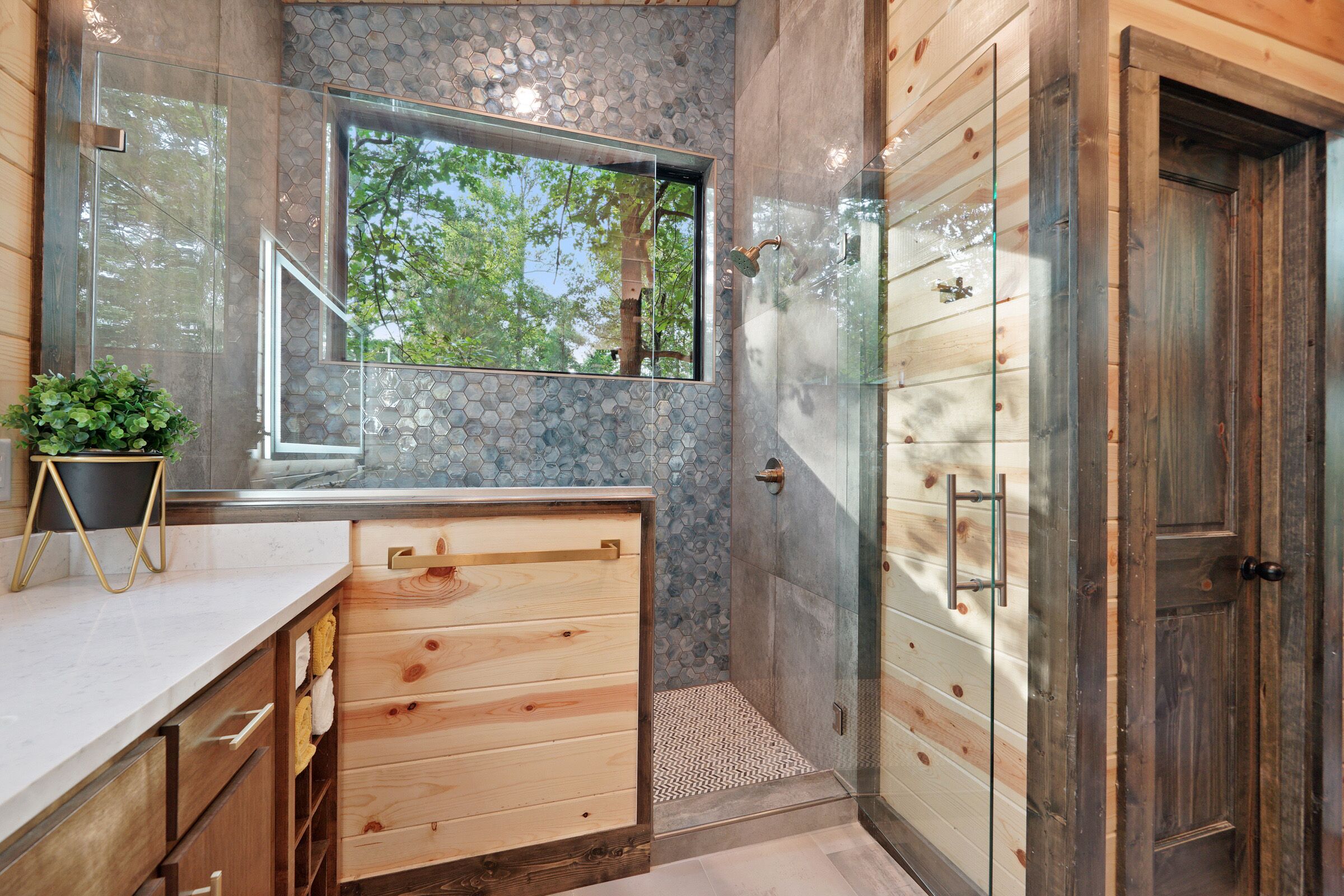 A modern bathroom with a wooden vanity, a glass shower enclosure, and a view of trees through a window.