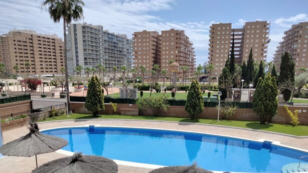 Una piscina con ombrelloni di paglia, circondata da vegetazione e alti edifici sullo sfondo.