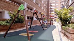Un'area giochi all'aperto con altalene e una struttura per arrampicarsi, circondata da un muro in mattoni e vegetazione.