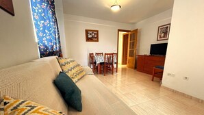 A living room with a sofa, patterned cushions, a dining table with chairs, a TV, and a wooden cabinet.