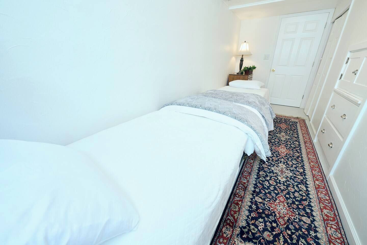 A narrow room with two single beds, a nightstand with a lamp, a door, and a patterned rug.